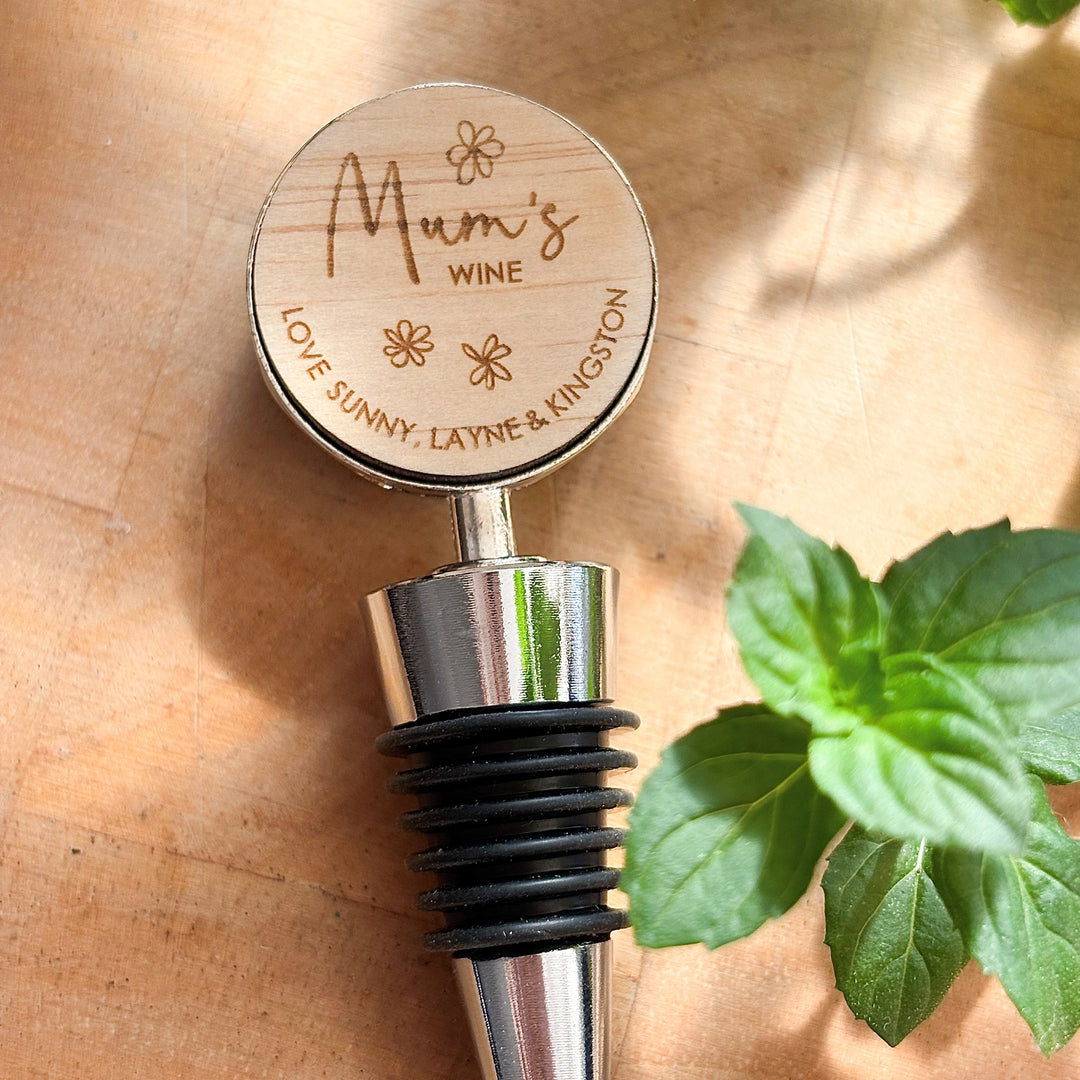 A personalised wine bottle stopper with engraved text on a wooden top, attached to a metallic stopper, placed on a wooden surface with a green leafy herb beside it.